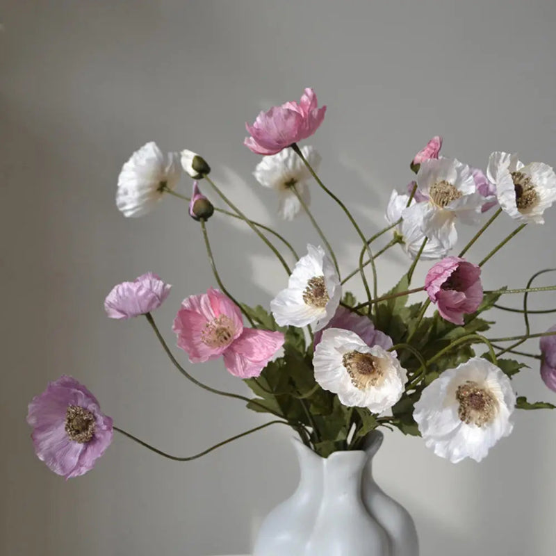 A white undulating vase holds a bouquet of white, pink, and purple poppies.