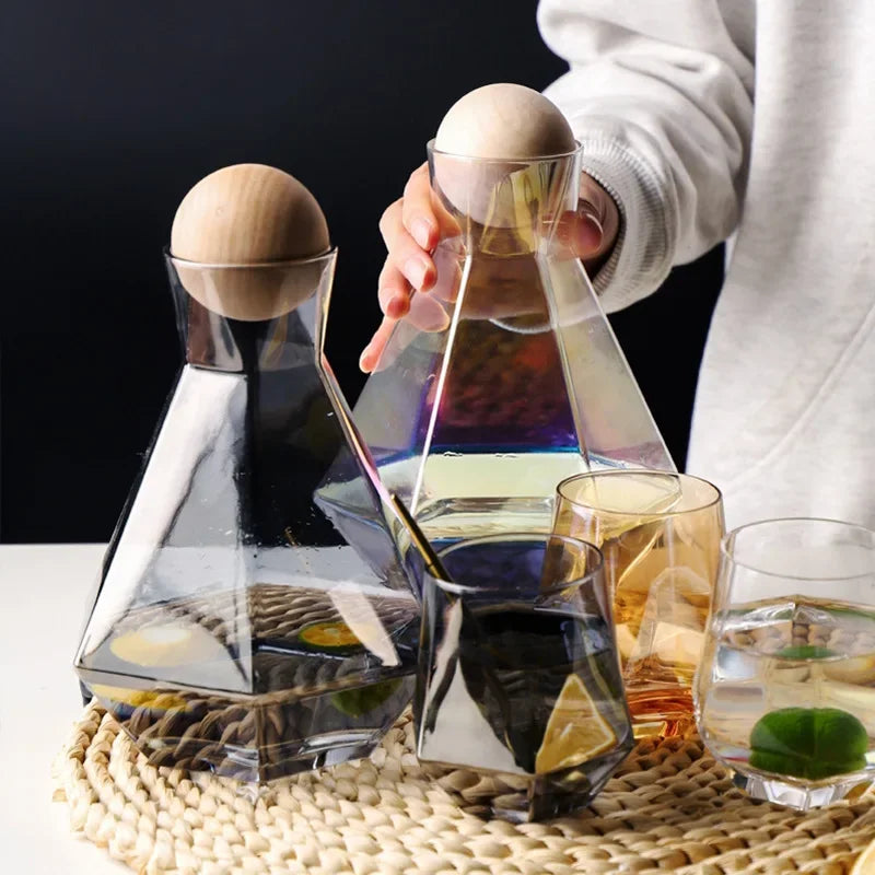 Two glass pitchers with wooden ball lids and matching faceted glasses, one clear and one iridescent, on a woven placemat with fruit slices in the drinks. A hand is reaching for the iridescent pitcher.