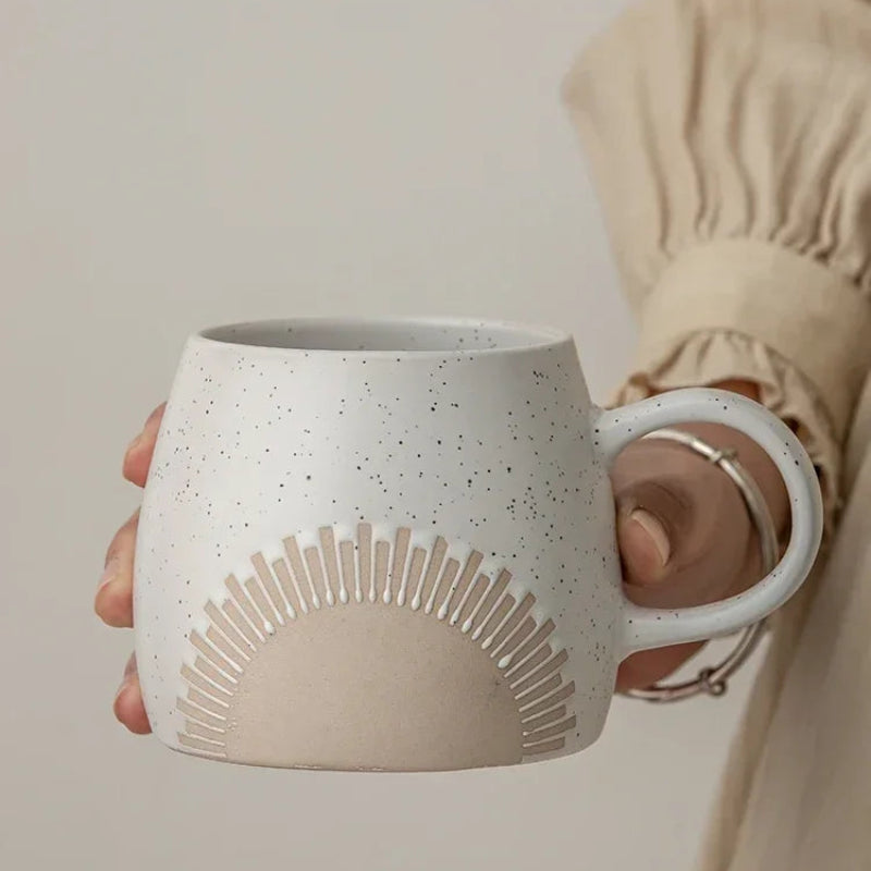A person wearing a beige, long-sleeved blouse holds a ceramic mug with a speckled matte glaze. The mug features an embossed sunburst design and a curved handle, offering a rustic yet modern aesthetic.