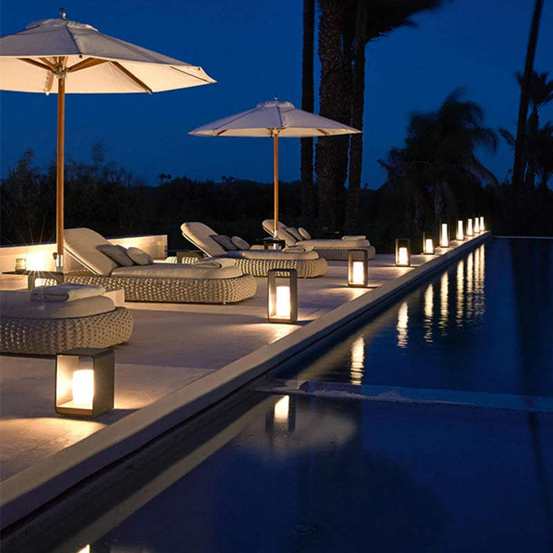 A tranquil nighttime scene of a poolside patio with lounge chairs under white umbrellas, illuminated by several modern, dark lanterns with warm glowing lights, reflecting in the calm water.