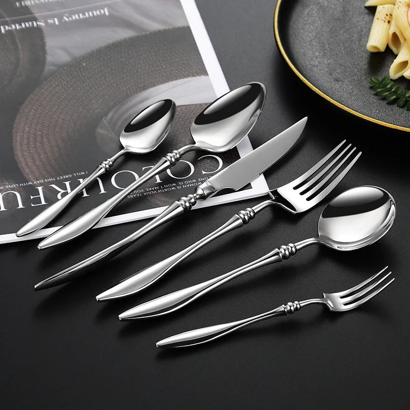 A set of six silver utensils with a magazine and food in the background.