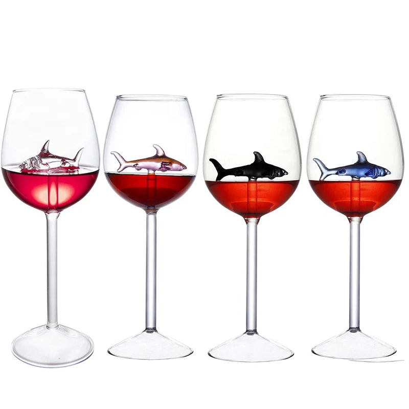 A product image showing four clear wine glasses, each with a different colored glass shark figurine suspended inside and filled with a red liquid.