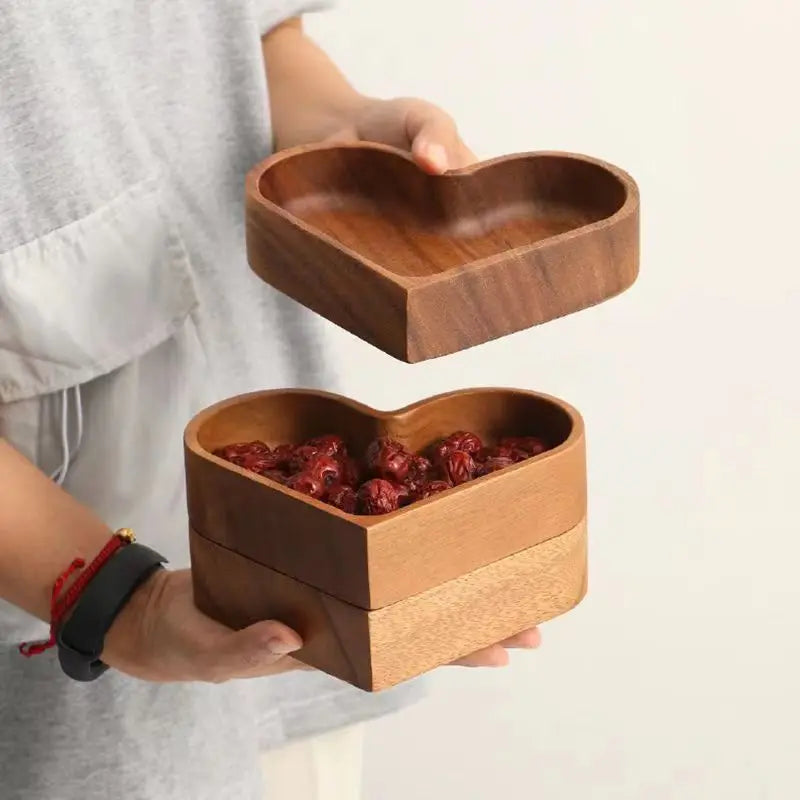 A person holding a heart-shaped wooden bowl filled with dried dates, with another empty heart-shaped wooden bowl floating above.