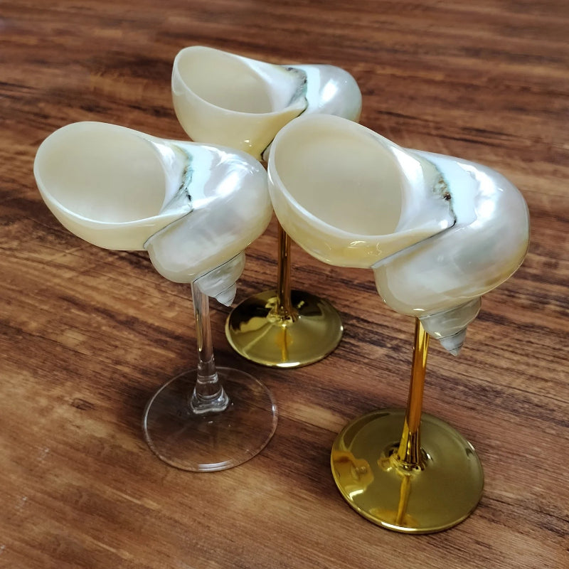 Three seashell wine glasses with clear or gold stems and a pearlescent seashell cup, arranged on a dark wooden surface.