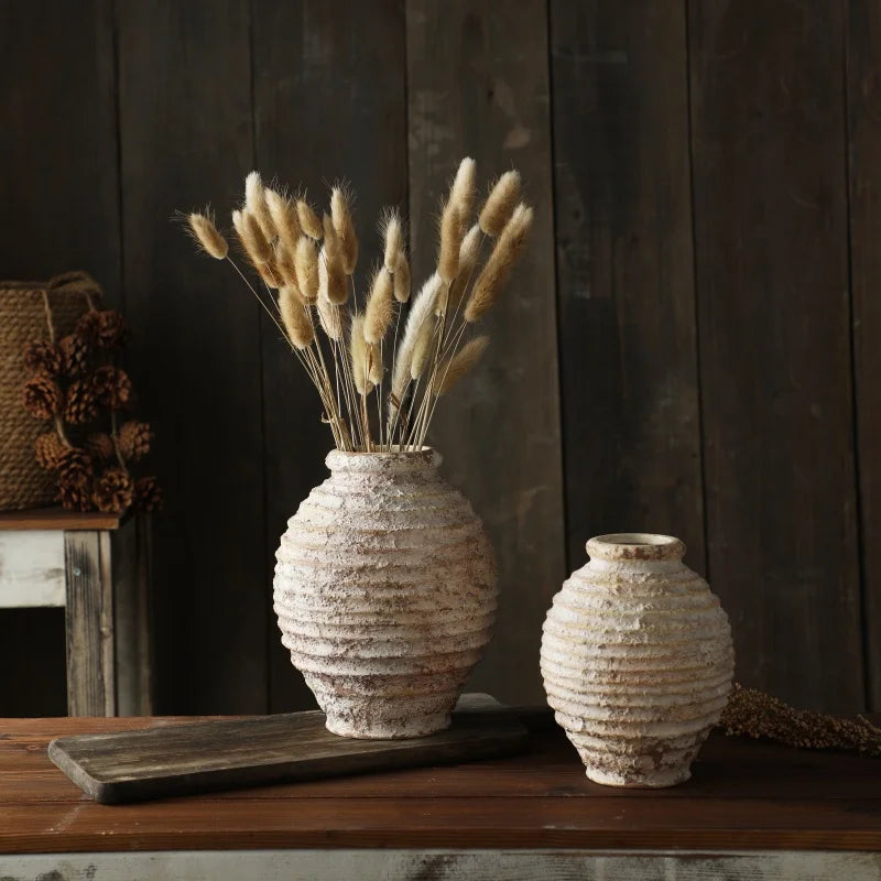 Two textured vases with dried plants on a wooden surface against a dark wooden wall.