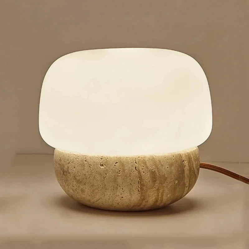 A small, glowing table lamp with a rounded white glass shade and a textured, light-colored stone base sits on a light surface. The lamp emits a soft, warm light, and its brown cord extends to the right. The background is a muted beige.