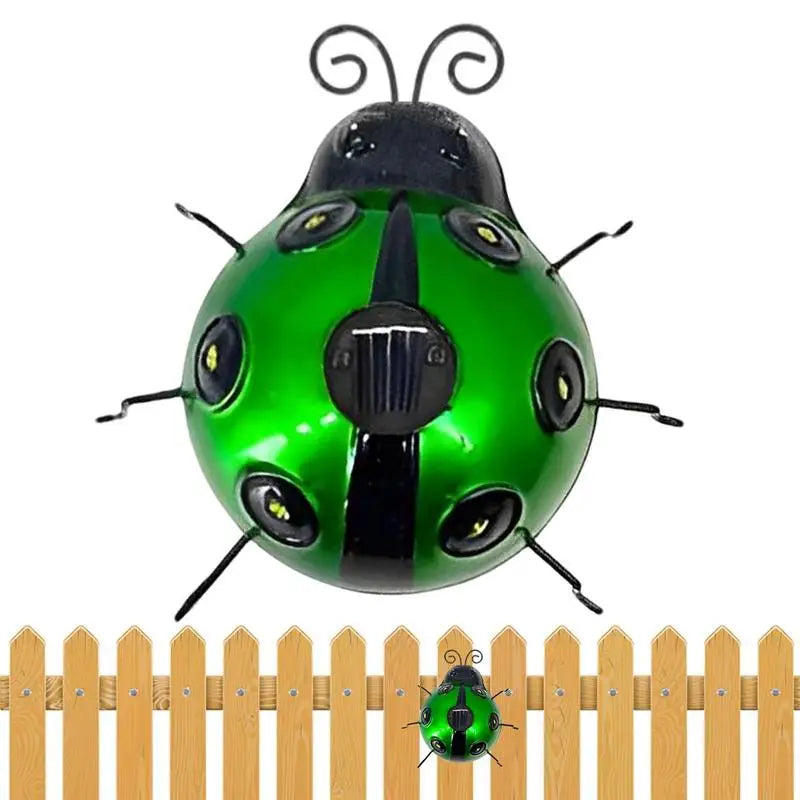 A decorative green and black metal ladybug with a small solar panel on its back is shown against a white background. It has black antennae and six black legs, with small lights embedded in its spots. A smaller version of the ladybug is shown in front of a short wooden fence at the bottom.