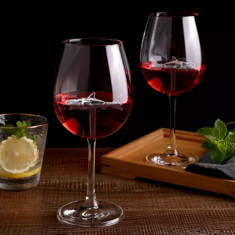 Two wine glasses filled with red wine, each containing a small, silver glass shark figurine. A blurry glass with a lemon slice is also on the table.