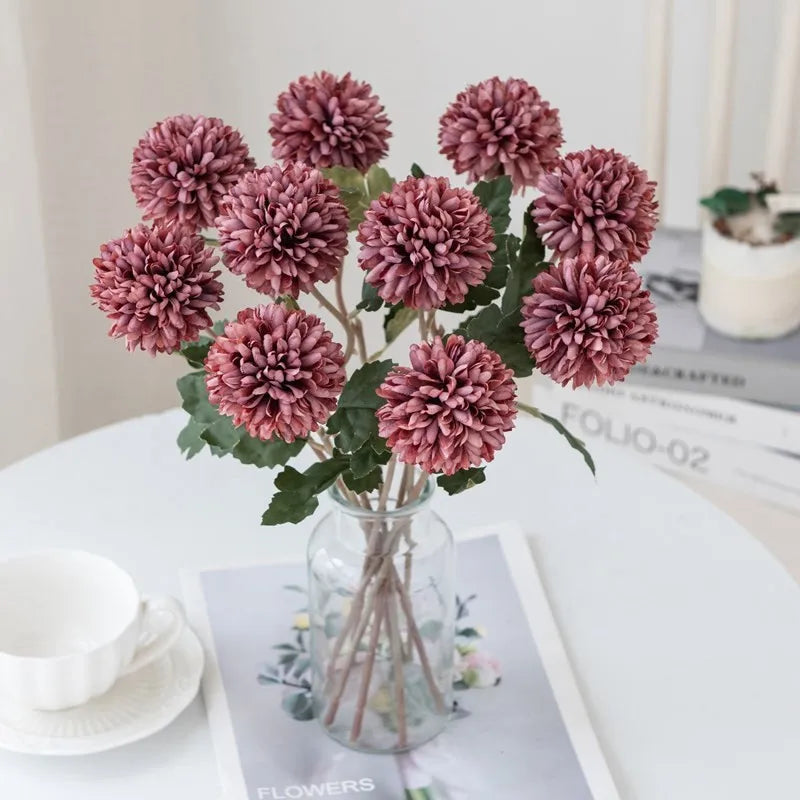 Maroon faux chrysanthemum flowers in a clear vase on a white table.