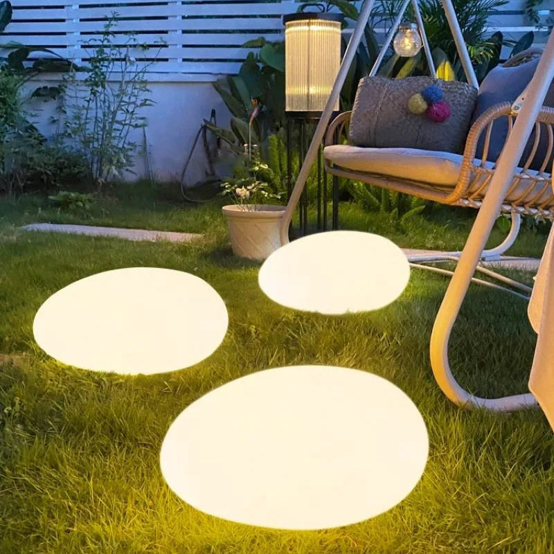 Three illuminated, round egg-shaped lights on a green lawn, with a hanging swing chair and standing lantern in the background.