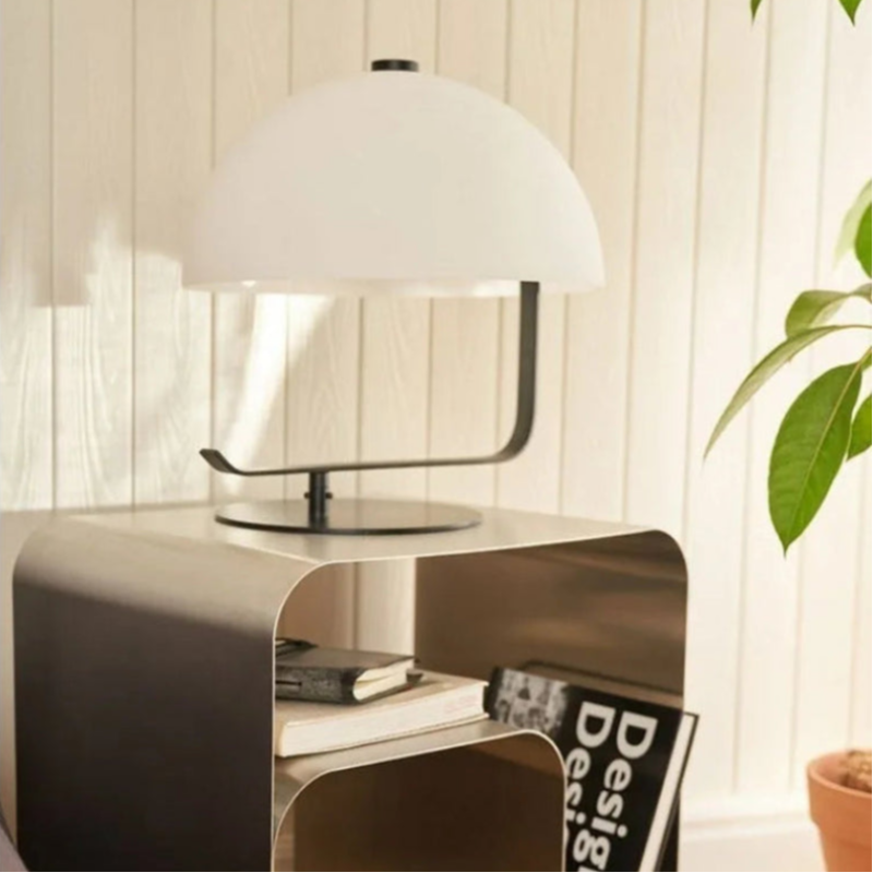 A half-dome shaped lamp with a black metal base and white shade sits on a silver, bent metal nightstand with rounded corners.