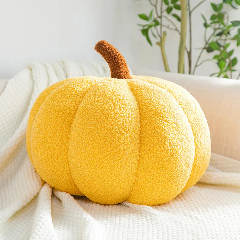 A plush, bright yellow pumpkin-shaped pillow with a brown textured stem rests on a white, textured blanket draped over a sofa.