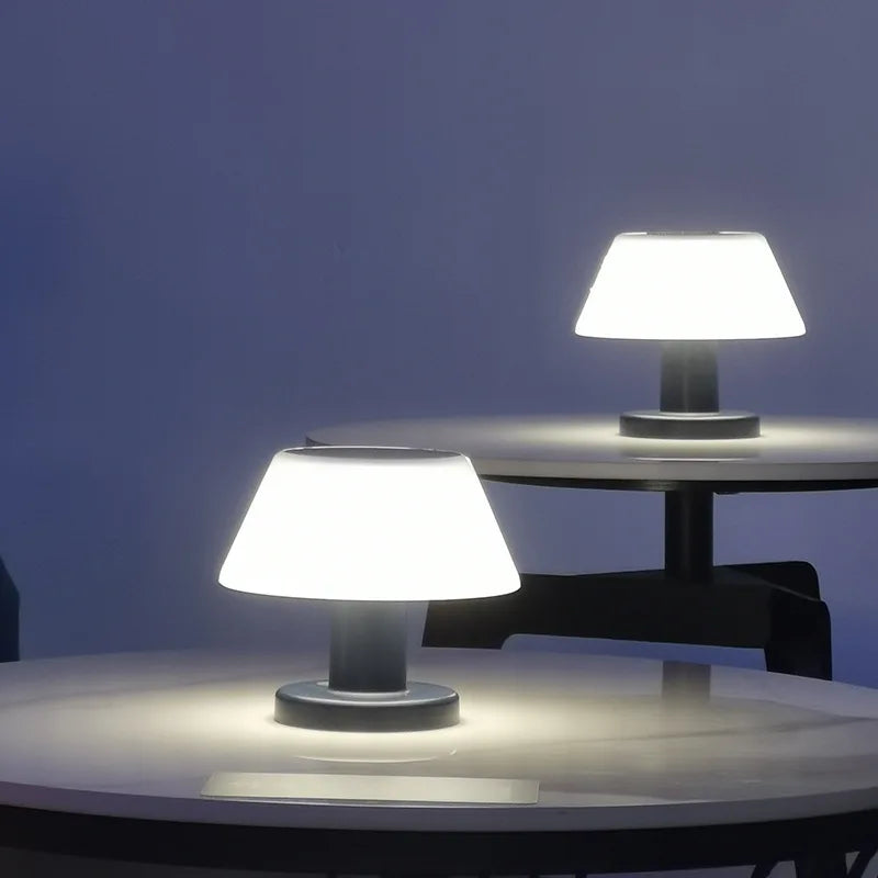 Two small, modern table lamps with white, slightly tapered shades and dark bases illuminate round tables in a dimly lit setting with a dark blue background. The lamps cast a soft, circular glow on the tabletops.