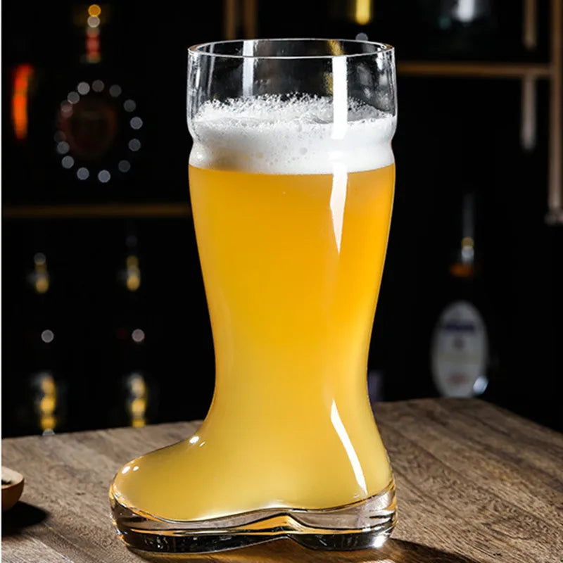 A clear glass beer boot mug filled with a light-colored beer and a thick white foam head sits on a wooden surface against a blurred background of a bar.