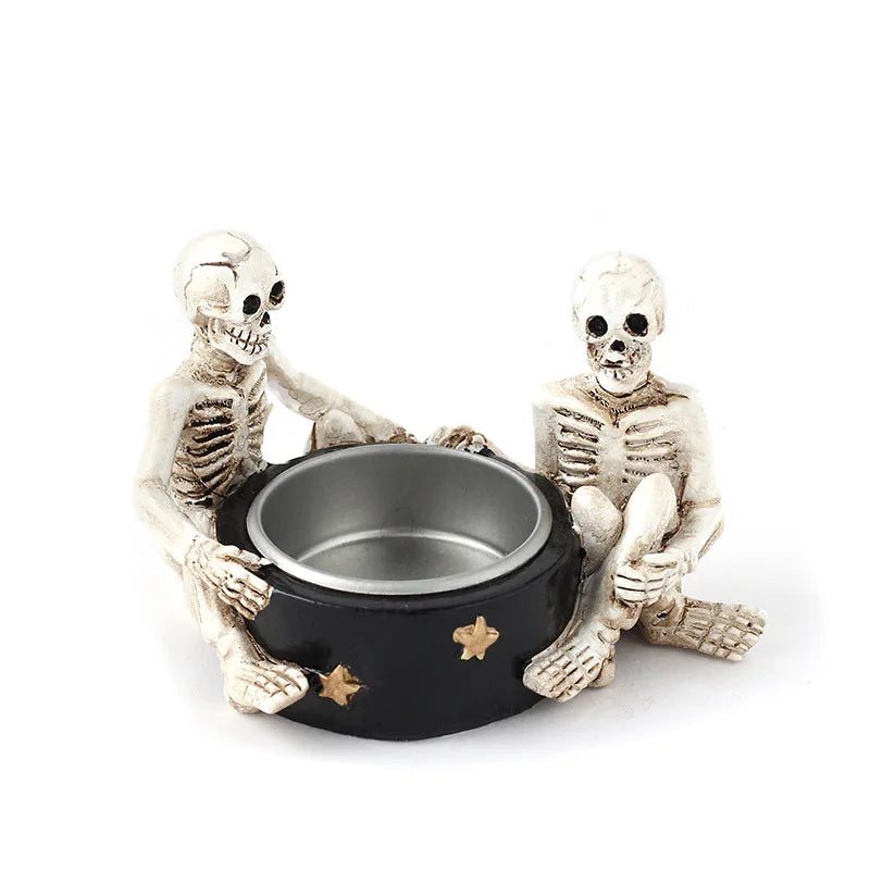 Two skeleton figurines sitting on either side of a black candle holder with gold stars, on a white background.