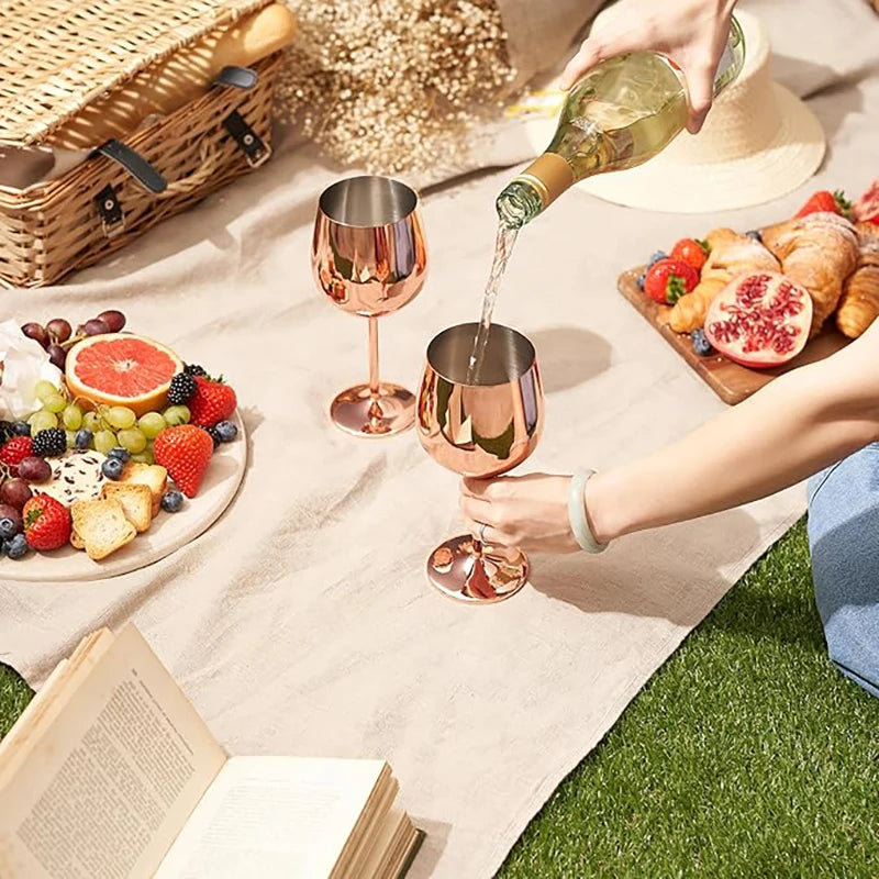 A picnic setting featuring two rose gold stainless steel wine glasses on a beige blanket. A person is pouring white wine into one of the glasses, while a spread of fresh fruits, croissants, and a pomegranate rests on wooden and ceramic serving boards. A woven picnic basket, a sun hat, and an open book complete the cozy outdoor ambiance.
