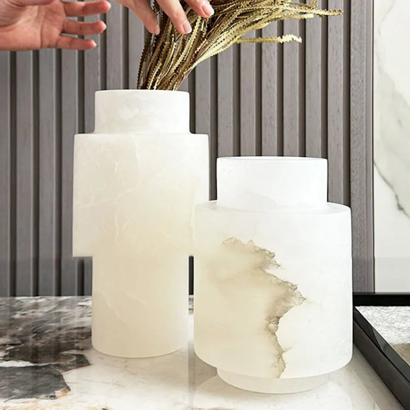 A pair of Scandinavian White Onyx vases displayed on a marble surface, featuring a modern geometric design with natural veining. One vase holds dried golden plants, highlighting its decorative use. The background showcases a contemporary interior with neutral tones.