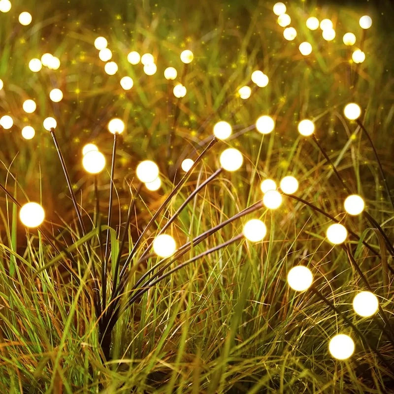 Numerous slender dark stems with glowing warm white spherical lights rise from tall green and brown grass in a garden at dusk or dawn. The lights are scattered throughout the grass, creating a magical, firefly-like ambiance.