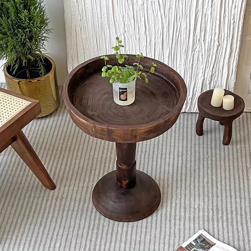 A rustic, dark wood, round side table (Momo Wabi-sabi Log Small Coffee Table) with a shallow tray top, holding a small potted plant, positioned on a striped rug with other decor elements.