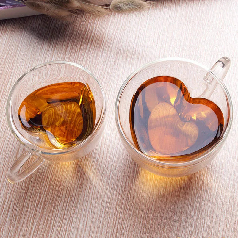 Two glass heart-shaped mugs filled with amber tea are on a wooden table.