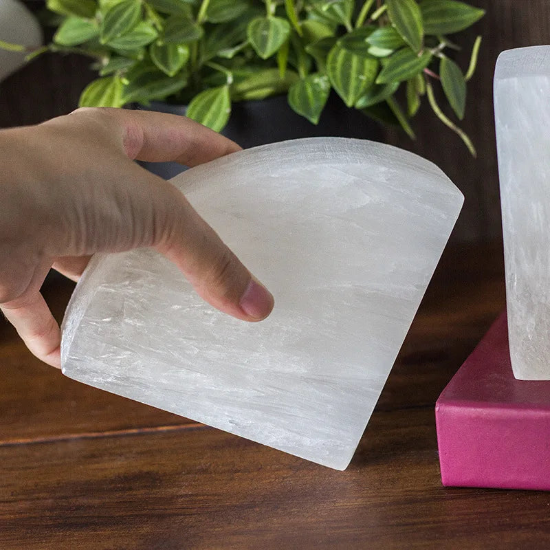 A person holding a white crystal bookend with a smooth, semi-circular shape, showcasing its size and texture. A wooden surface, a red book, and green plants are in the background.