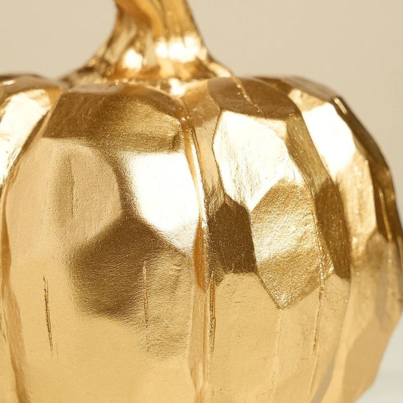 A close-up of a small, decorative golden pumpkin. The surface has a faceted, geometric texture that reflects light, and there is a visible seam or imperfection running down the front. The pumpkin's stem is visible at the top.