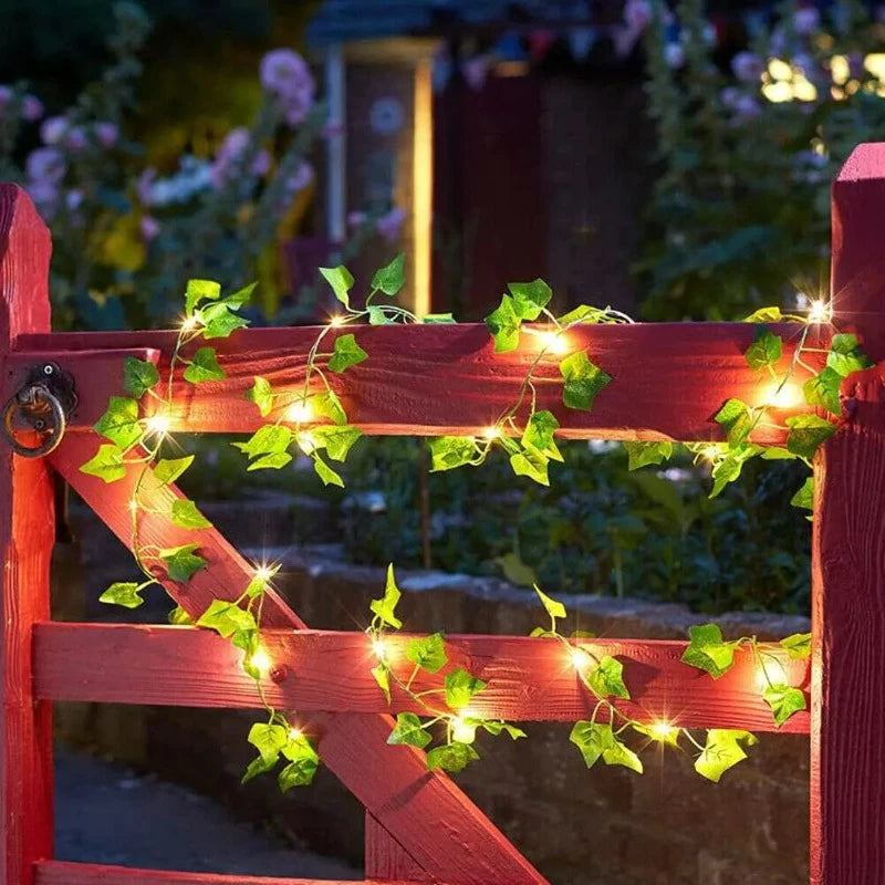 A string of warm white LED lights intertwined with artificial green ivy leaves is wrapped around a red wooden gate at night. The lights illuminate the gate and the surrounding foliage.
