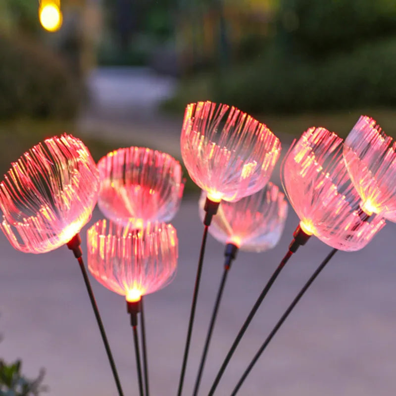 Several solar-powered garden lights with a translucent, jellyfish-like shape emit a soft pink glow at dusk or night. They are on thin dark stems and are seen outdoors with a blurred background.