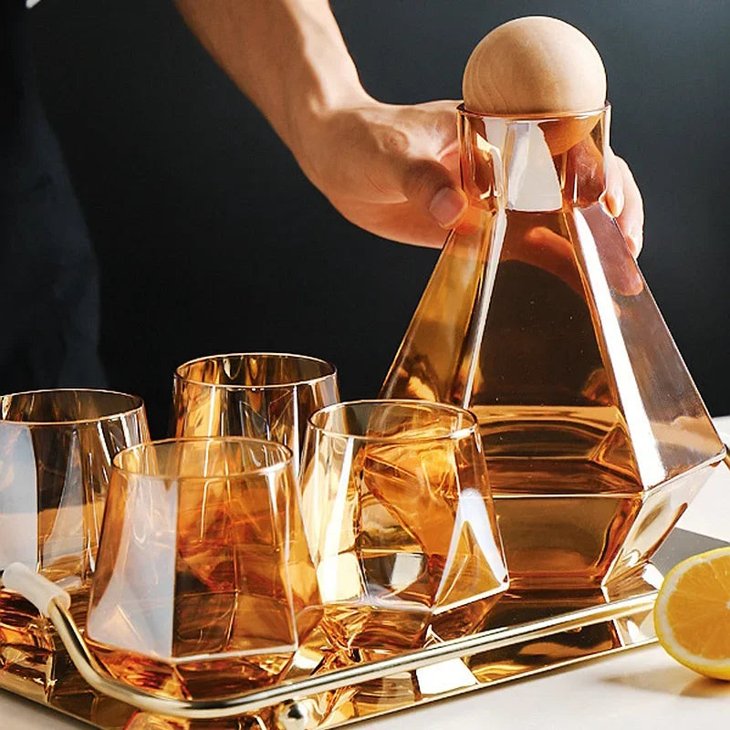 Amber glass pitcher and four matching faceted glasses on a gold tray. A hand is reaching towards the pitcher. A lemon slice is visible on the edge of the tray.