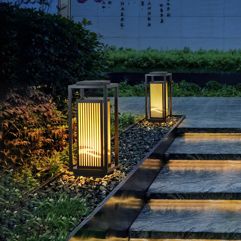 Two modern, dark-framed outdoor lanterns with vertical slats emit a warm glow along a stone pathway with steps at dusk or dawn. The lanterns sit on dark gravel beside the illuminated walkway, with lush greenery and a wall with script in the background.
