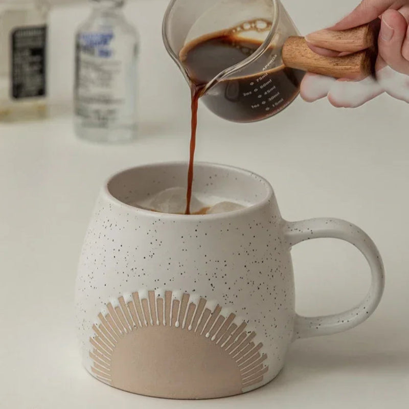 A speckled ceramic mug with an embossed sunburst design is filled with ice as a hand pours freshly brewed coffee from a glass beaker with a wooden handle. The minimalist background features bottles and a clean, neutral-toned surface.