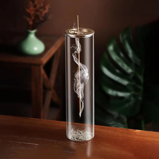 A sleek and modern incense burner featuring a transparent glass cylinder with a suspended incense stick inside. The smoke swirls elegantly as it rises, creating a calming visual effect. The ash collects neatly at the bottom, maintaining a clean setup. The background includes a wooden table, a green vase with dried flowers, and a large tropical leaf, enhancing the serene and minimalist ambiance.