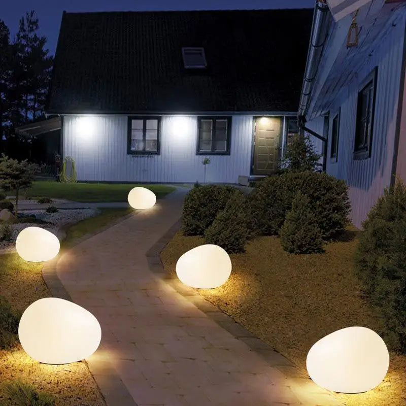 A nighttime view of five large, round, illuminated lights placed along a stone pathway leading to a two-story house with a dark roof. The house has a door and windows visible, and the lights provide a warm glow against the dark surroundings.