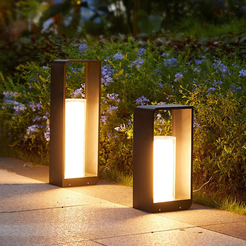 Two modern, dark rectangular outdoor pathway lights with tall, glowing cylindrical diffusers, illuminating a stone walkway bordered by lush greenery and small blue flowers at dusk.