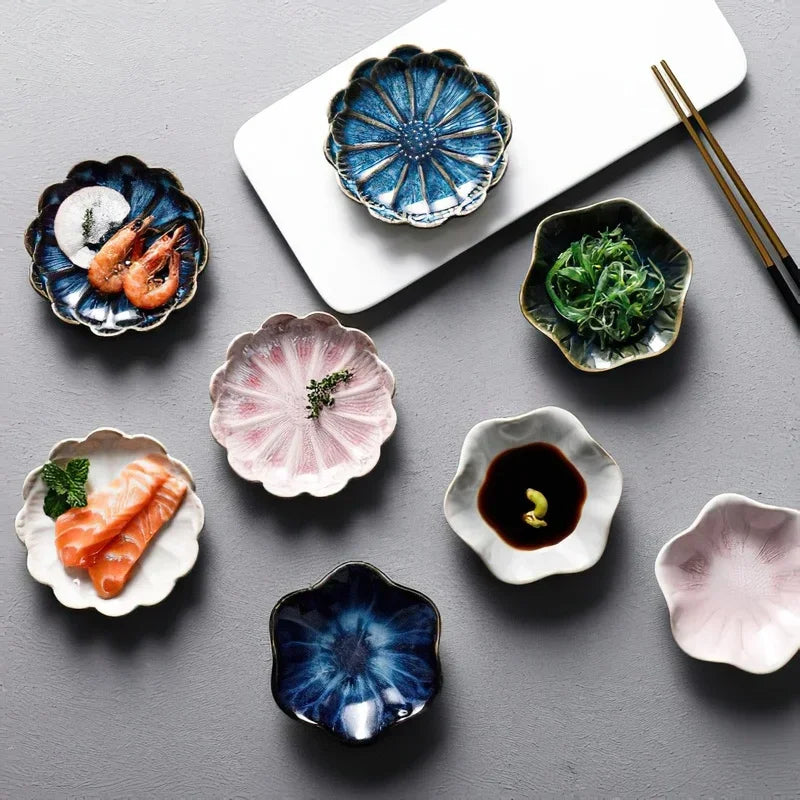 Small, flower-shaped dishes holding various foods like shrimp, salmon, seaweed, and soy sauce, scattered on a gray surface next to a white serving board and chopsticks.
