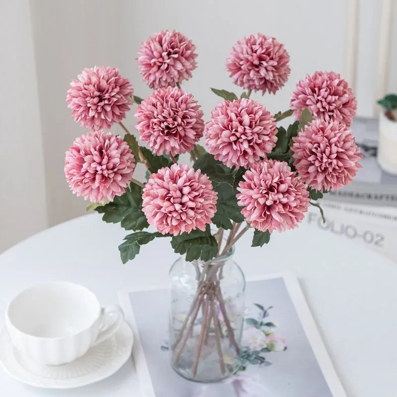 Pink faux chrysanthemum flowers in a clear vase on a white table.