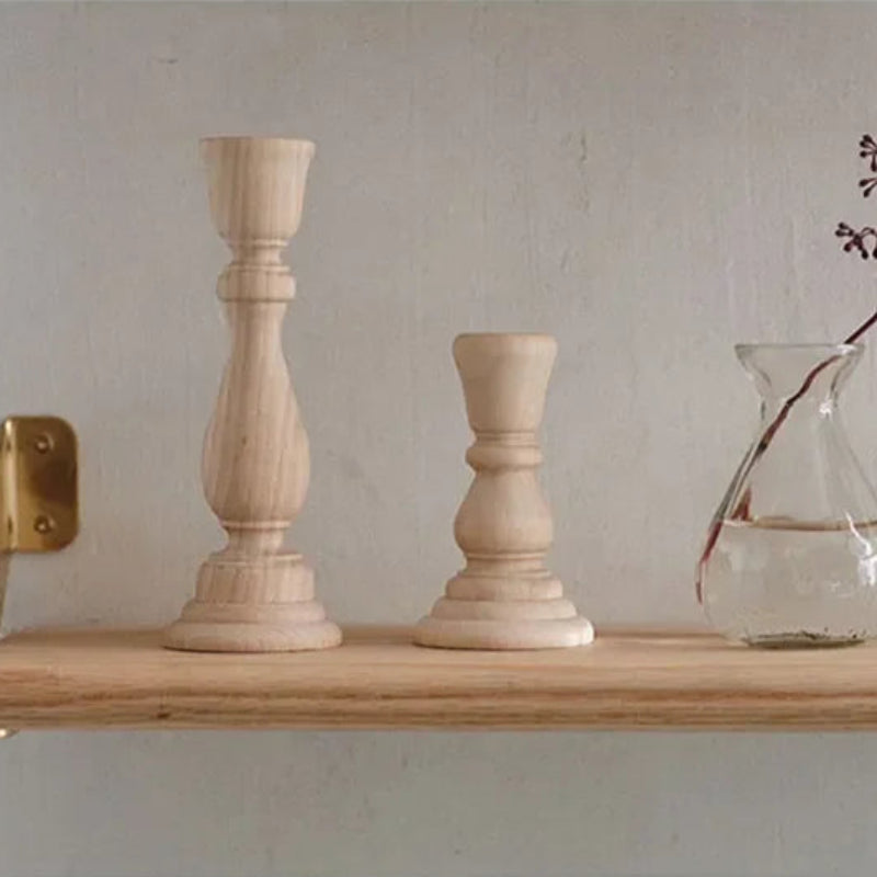Two unvarnished, light wood candlestick holders, one tall and one short, sit on a wood shelf next to a small glass vase with a plant