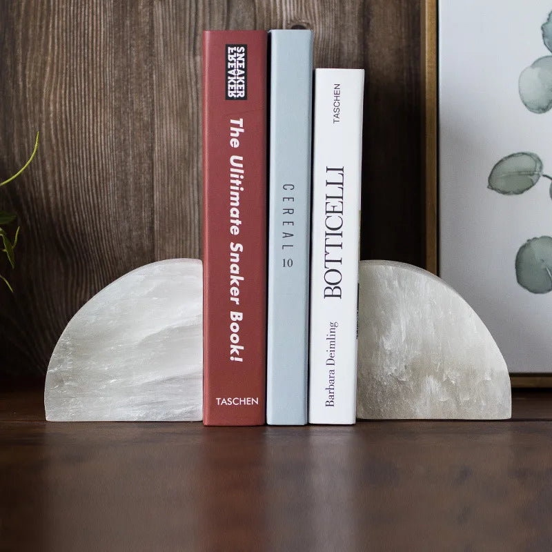 A pair of white crystal bookends with a smooth, semi-circular design holding up three books on a wooden surface. The background features a wooden wall, a framed botanical print, and a small potted plant.