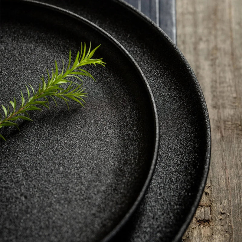 A close-up of two speckled black plates on a rustic wooden table, with a green sprig on the top plate.