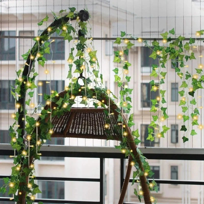 A hanging wicker swing chair on a balcony is decorated with artificial green ivy vines intertwined with warm white fairy lights. The lights and foliage are draped along the chair's frame and hang down in strands. Tall buildings are visible in the blurred background through a mesh railing.