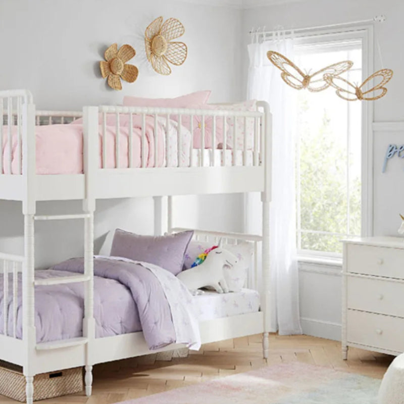 A cozy children's bedroom featuring a white wooden bunk bed with pastel-colored bedding—soft pink on the top bunk and lavender on the bottom. A plush unicorn toy rests on the lower bed. The room is decorated with woven rattan flower wall hangings above the bed and delicate rattan butterfly wall art near a large window with sheer white curtains. A white dresser sits in the corner, adding to the light and airy aesthetic of the space.
