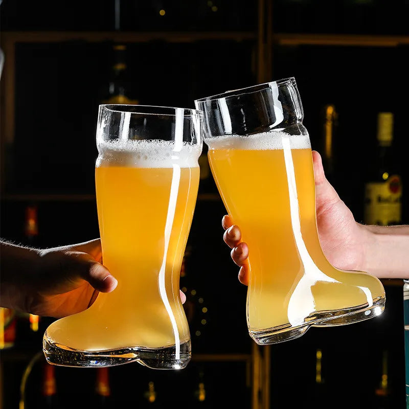 Two hands holding clear glass beer boot mugs filled with light-colored beer and white foam, clinking them together in a dimly lit bar setting with blurred shelves of bottles in the background.
