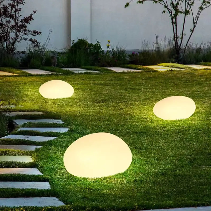 Three illuminated, round egg-shaped lights scattered across a green lawn with a stone pathway winding through it.