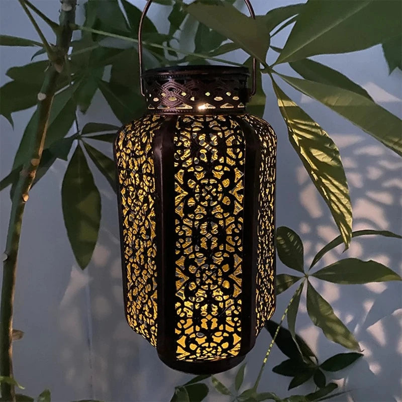 A decorative metal solar lantern with intricate cutouts hangs from a branch, emitting a warm glow.