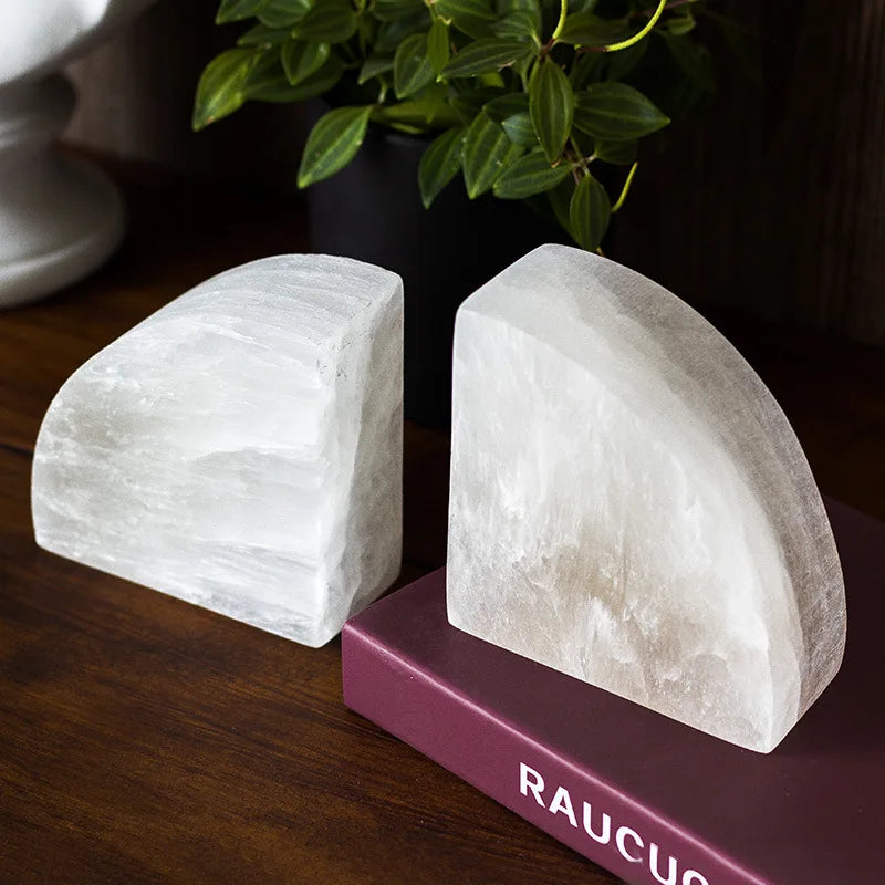 A pair of white crystal bookends with a smooth, semi-circular design displayed on a wooden surface. One bookend rests on a red book, while a green potted plant adds a decorative touch in the background.