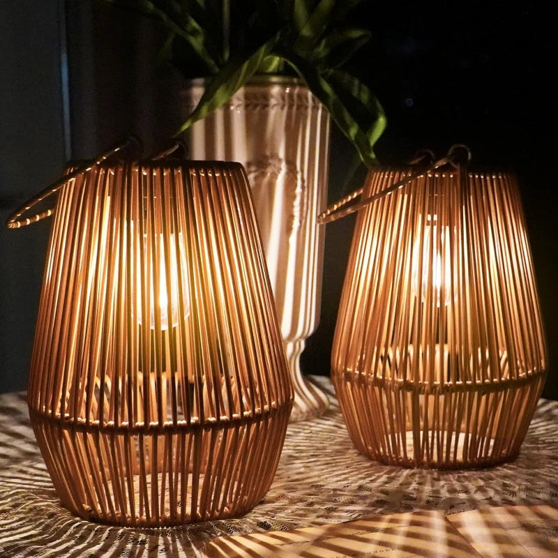 Two warm-toned, cylindrical outdoor lanterns made of vertical slats emit a soft glow as they sit on a surface with a striped pattern. A light-colored vase with greenery is blurred in the background. The lanterns have handles for hanging.