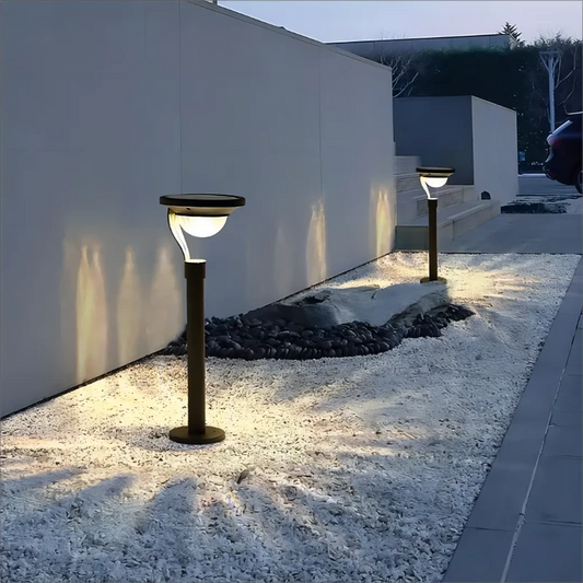 Two modern black solar pathway lights with curved tops illuminate a walkway covered in small white stones at dusk or night. The lights cast a warm downward glow. A light-colored wall runs along the left side.
