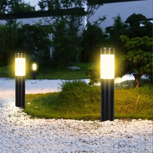 Three cylindrical outdoor pathway lights illuminate a garden area with white gravel, patches of grass, and various plants at night. The lights have dark cylindrical bases and frosted white diffusers, emitting a warm, upward glow. The lights are positioned to illuminate the pathway and surrounding greenery. Trees and a dark sky are visible in the background.