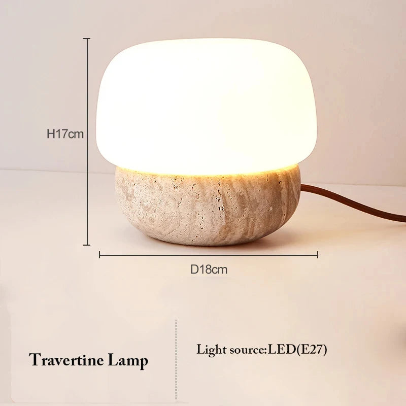 A small illuminated table lamp with a rounded white shade and a textured, light-colored travertine base is shown against a light background with dimensional measurements and light source information. The lamp has a height (H) of 17cm and a diameter (D) of 18cm. Text indicates it is a "Travertine Lamp" with an "LED (E27)" light source. A brown power cord extends to the right.
