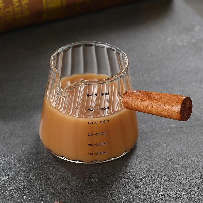 Glass measuring cup with a ribbed design, a wooden handle, and a small spout, filled with coffee or tea. Measurement markings in ounces and milliliters are visible on the side. The cup sits on a textured countertop with a coffee bean nearby.